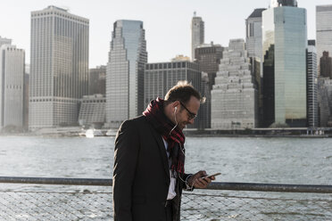 USA, Brooklyn, businessman with smartphone and earphones in front of Manhattan skyline - UUF09285