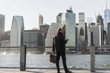 USA, Brooklyn, businessman with briefcase and smartphone standing in front of Manhattan skyline - UUF09287