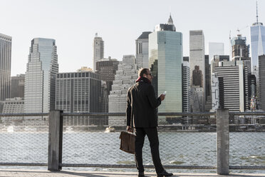 USA, Brooklyn, Geschäftsmann mit Aktentasche und Smartphone vor der Skyline von Manhattan stehend - UUF09287