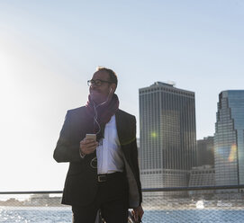 USA, Brooklyn, smiling businessman with earphones and smartphone in front of Manhattan skyline - UUF09288