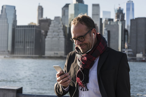 USA, Brooklyn, lächelnder Geschäftsmann mit Kopfhörern, der auf sein Smartphone schaut, lizenzfreies Stockfoto