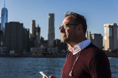 USA, Brooklyn, Geschäftsmann mit Smartphone und Kopfhörern im Sonnenlicht, lizenzfreies Stockfoto