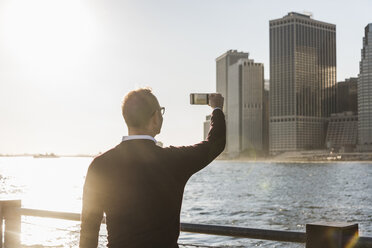 USA, Brooklyn, man taking picture of Manhattan skyscrapers with smartphone at evening twilight - UUF09294