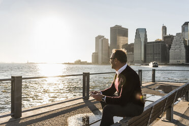 USA, Brooklyn, entspannter Mann mit Smartphone, der auf einer Bank sitzt und die Aussicht betrachtet - UUF09296