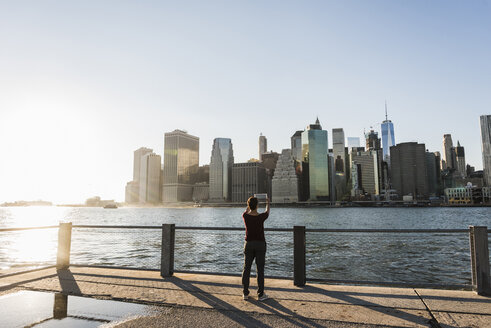 USA, Brooklyn, Rückenansicht einer Frau, die mit einem Tablet die Skyline von Manhattan fotografiert - UUF09302