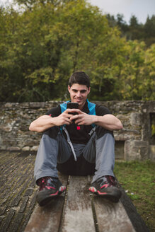 Lächelnder Wanderer, der auf einer Bank sitzt und in der Natur auf sein Smartphone schaut - RAEF01570