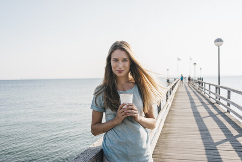 Porträt einer glücklichen jungen Frau mit Kaffee zum Mitnehmen auf dem Bootssteg - KNSF00672