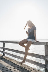 Junge Frau steht auf einem Steg im Gegenlicht und schaut auf ihr Handy - KNSF00677