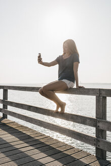 Junge Frau sitzt auf dem Steg im Gegenlicht und macht ein Selfie mit ihrem Handy - KNSF00678