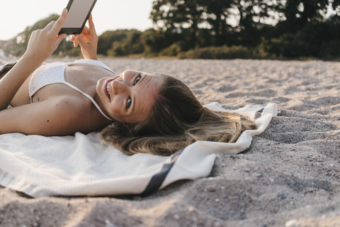 Lächelnde junge Frau auf Decke am Strand mit Tablet liegend, lizenzfreies Stockfoto