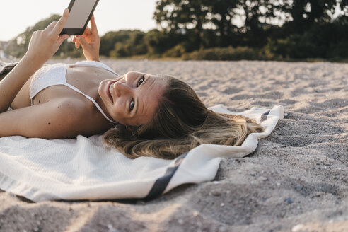 Lächelnde junge Frau auf Decke am Strand mit Tablet liegend - KNSF00688