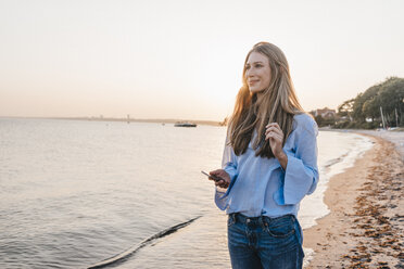 Smiling young woman lwith smartphone standing on the beach - KNSF00699