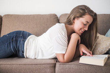 Lächelnde junge Frau liegt auf der Couch und liest ein Buch - SIPF01120