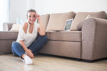 Smiling young woman sitting on the floor of living room listening music with laptop and headphones - SIPF01136