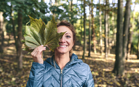 Grinsende Frau, die ihr Auge mit einem Herbstblatt im Wald bedeckt, lizenzfreies Stockfoto