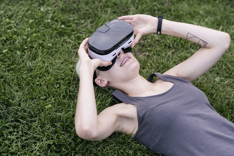 Junge Frau liegt auf einer Wiese und trägt eine Virtual-Reality-Brille im Freien, lizenzfreies Stockfoto