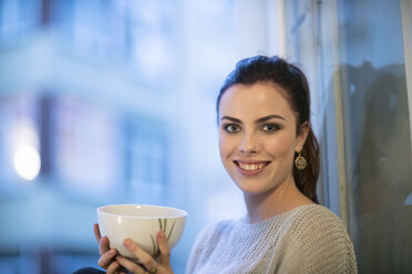Portrait of smiling young woman with tea bowl in front of window - TAMF00843
