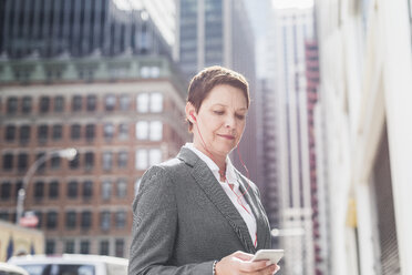 USA, New York City, Geschäftsfrau in Manhattan mit Kopfhörern, die auf ein Mobiltelefon schaut - UUF09404