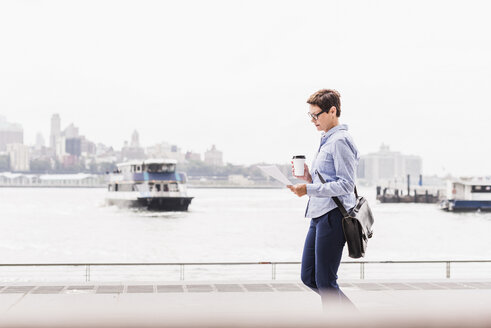 USA, New York City, Geschäftsfrau, die am East River spazieren geht und ein Dokument liest - UUF09413