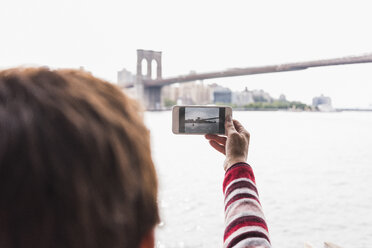 USA, New York City, Frau am East River, die mit ihrem Handy ein Foto von der Brooklyn Bridge macht - UUF09428
