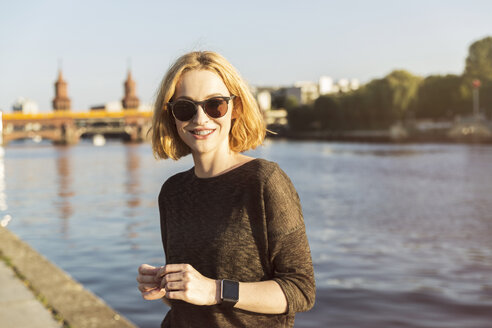 Deutschland, Berlin, Porträt einer lächelnden jungen Frau mit Sonnenbrille und Smartwatch - TAMF00858