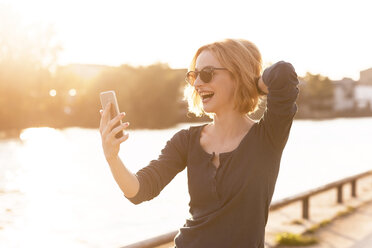 Glückliche junge Frau nimmt Selfie mit Smartphone bei Gegenlicht - TAMF00862