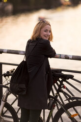 Porträt einer lächelnden jungen Frau mit Fahrrad, die sich in der Abenddämmerung an ein Geländer lehnt - TAMF00877