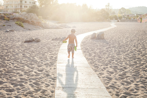 Rückansicht eines kleinen Jungen, der bei Gegenlicht auf der Strandpromenade läuft, lizenzfreies Stockfoto