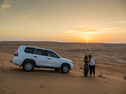 Oman, Al Raka, zwei junge Frauen stehen neben einem Geländewagen auf einer Wüstendüne und beobachten den Sonnenuntergang - AMF05096