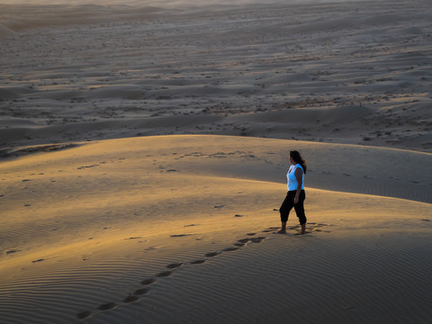 Oman, Al Raka, junge Frau steht auf einer Wüstendüne in der Wüste Rimal Al Wahiba und beobachtet den Sonnenuntergang, lizenzfreies Stockfoto