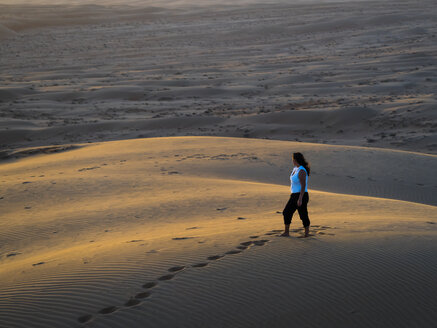 Oman, Al Raka, young woman standing on a desert dune in Rimal Al Wahiba desert watching sunset - AMF05098