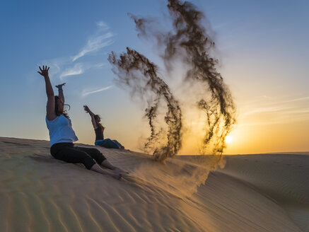 Oman, Al Raka, zwei junge Frauen sitzen auf einer Düne in der Wüste Rimal Al Wahiba und werfen Sand in die Luft - AMF05101