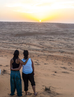 Oman, Al Raka, zwei junge Frauen stehen Arm in Arm auf einer Wüstendüne und beobachten den Sonnenuntergang - AMF05103