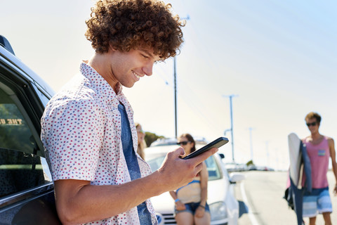 Lächelnder junger Mann an einem Auto, der sein Mobiltelefon überprüft, lizenzfreies Stockfoto