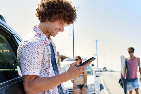 Lächelnder junger Mann an einem Auto, der sein Mobiltelefon überprüft - WESTF21979