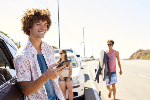 Lächelnder junger Mann an einem Auto mit Mobiltelefon in der Hand - WESTF21981