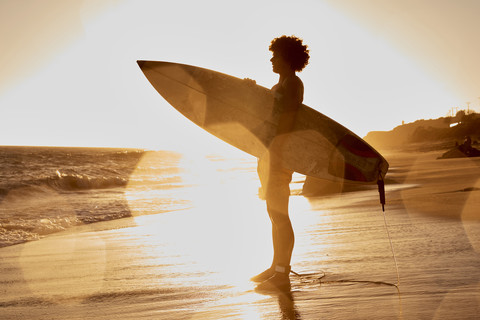 Junger Mann mit Surfbrett am Strand bei Sonnenuntergang, lizenzfreies Stockfoto