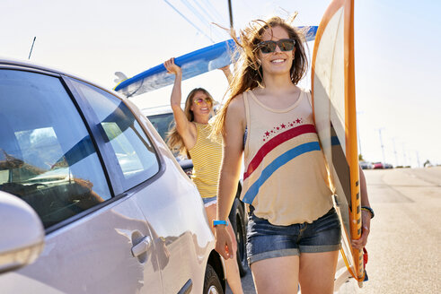 Two smiling young women carrying surfboards on coastal road - WESTF21995