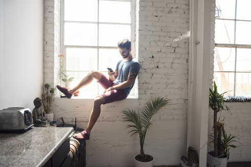 Junger Mann sitzt auf der Fensterbank und schaut auf sein Handy in einem Loft - WESTF22082