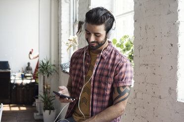 Smiling young man wearing headphones looking on cell phone in a loft - WESTF22088