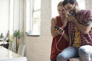 Glückliches junges Paar mit Mobiltelefon am Fenster, das sich Kopfhörer teilt - WESTF22104