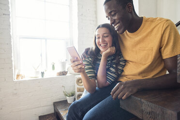 Happy young couple sitting on stairs in a loft sharing cell phone - WESTF22126