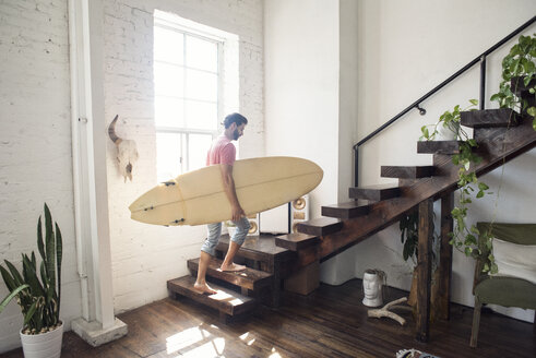 Young man carrying a surfboard on stairs in a loft - WESTF22129