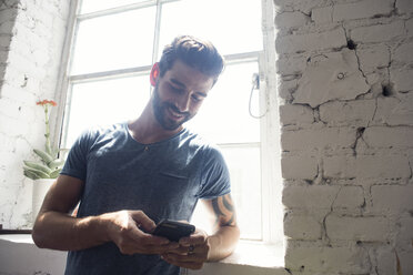 Lächelnder junger Mann, der am Fenster auf sein Handy schaut - WESTF22139