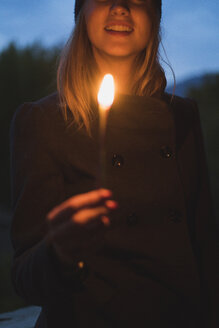 Lächelnde junge Frau, die in der Abenddämmerung ein Licht anzündet - KKAF00106