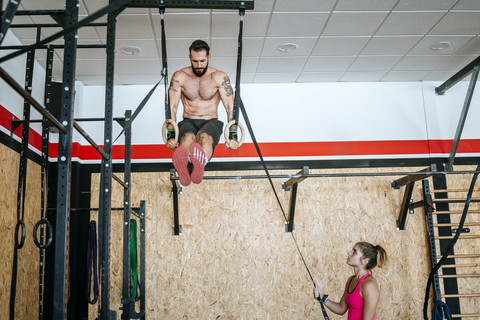 Mann macht Übungen an Ringen im Fitnessstudio mit Frau, die ihm hilft, lizenzfreies Stockfoto
