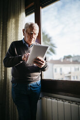 Senior man with tablet standing in front of window at home - JRFF01061