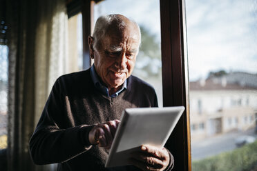 Senior man with tablet standing in front of window at home - JRFF01062