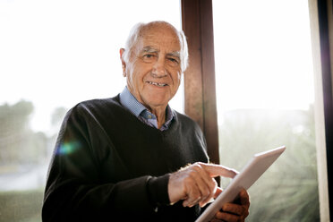 Portrait of smiling senior man with tablet standing in front of window - JRFF01064