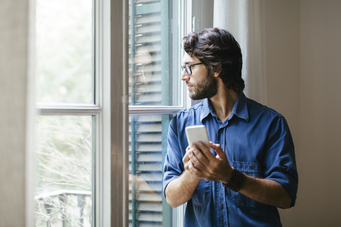 Geschäftsmann steht am Bürofenster und benutzt ein Smartphone, lizenzfreies Stockfoto